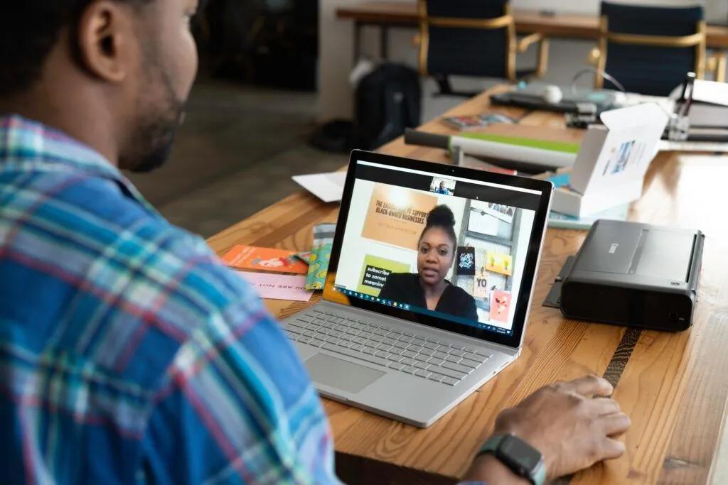 A man in a plaid shirt on a video call on his laptop at a wooden desk, with a woman visible on the screen