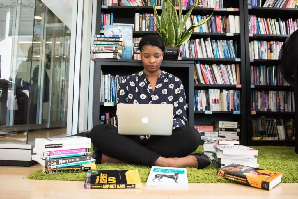 A woman sitting cross-legged on the floor using a laptop, surrounded by stacks of books in front of large bookshelves